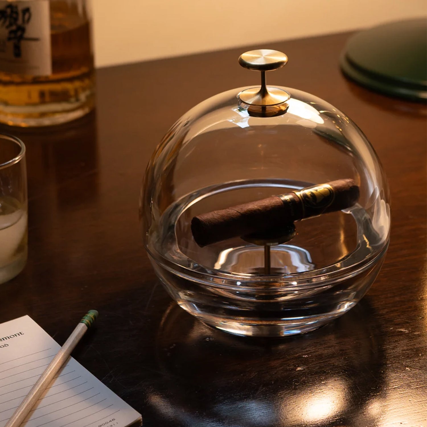 A clear glass dome cigar ashtray with a brass knob lid sits on a dark wooden table, containing a dark brown cigar. A glass of whiskey, a notebook with a pencil, and another green object are blurred in the background.