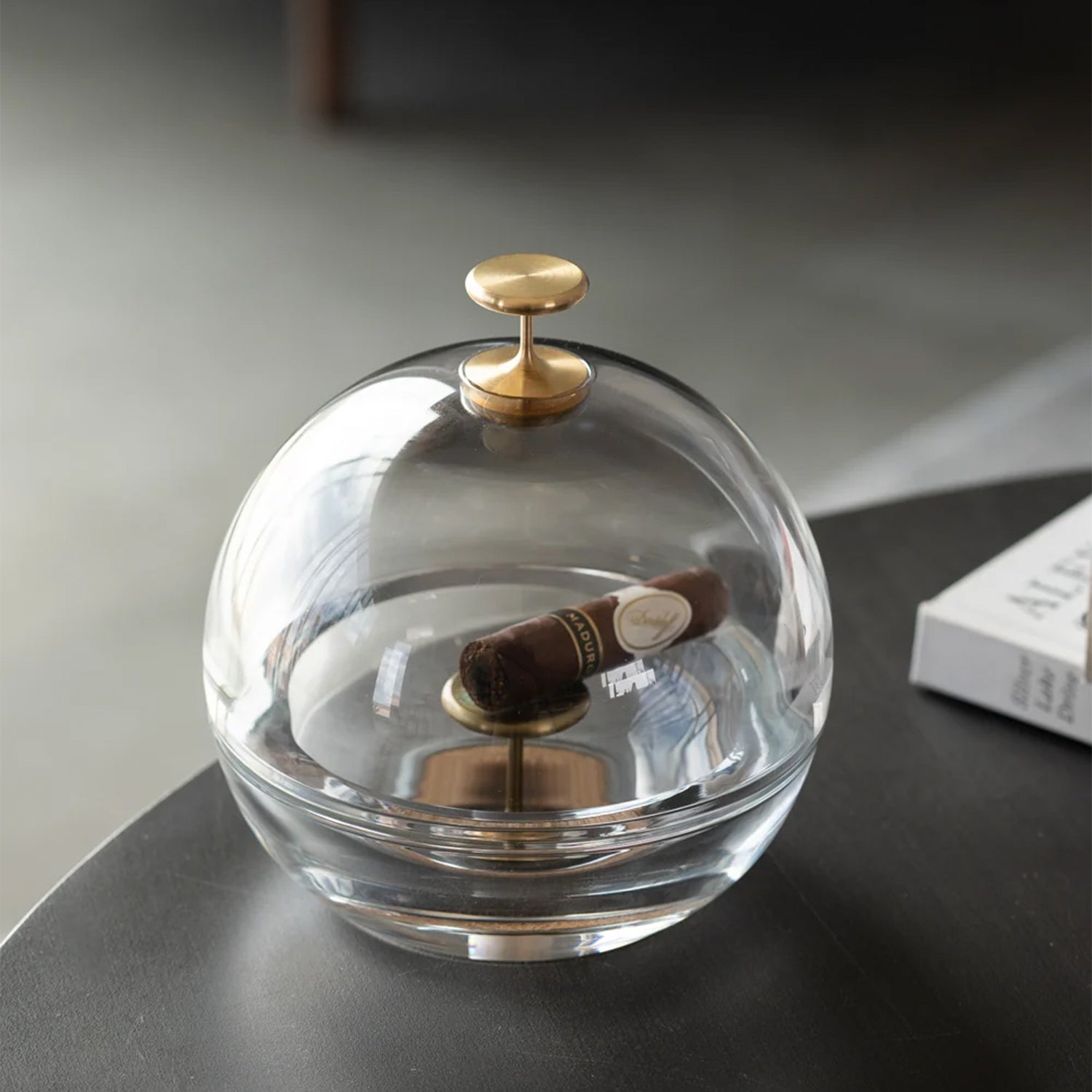 A clear glass dome-shaped cigar ashtray with a round top and polished gold-tone metal accents, shown open with a cigar inside on a dark tabletop.