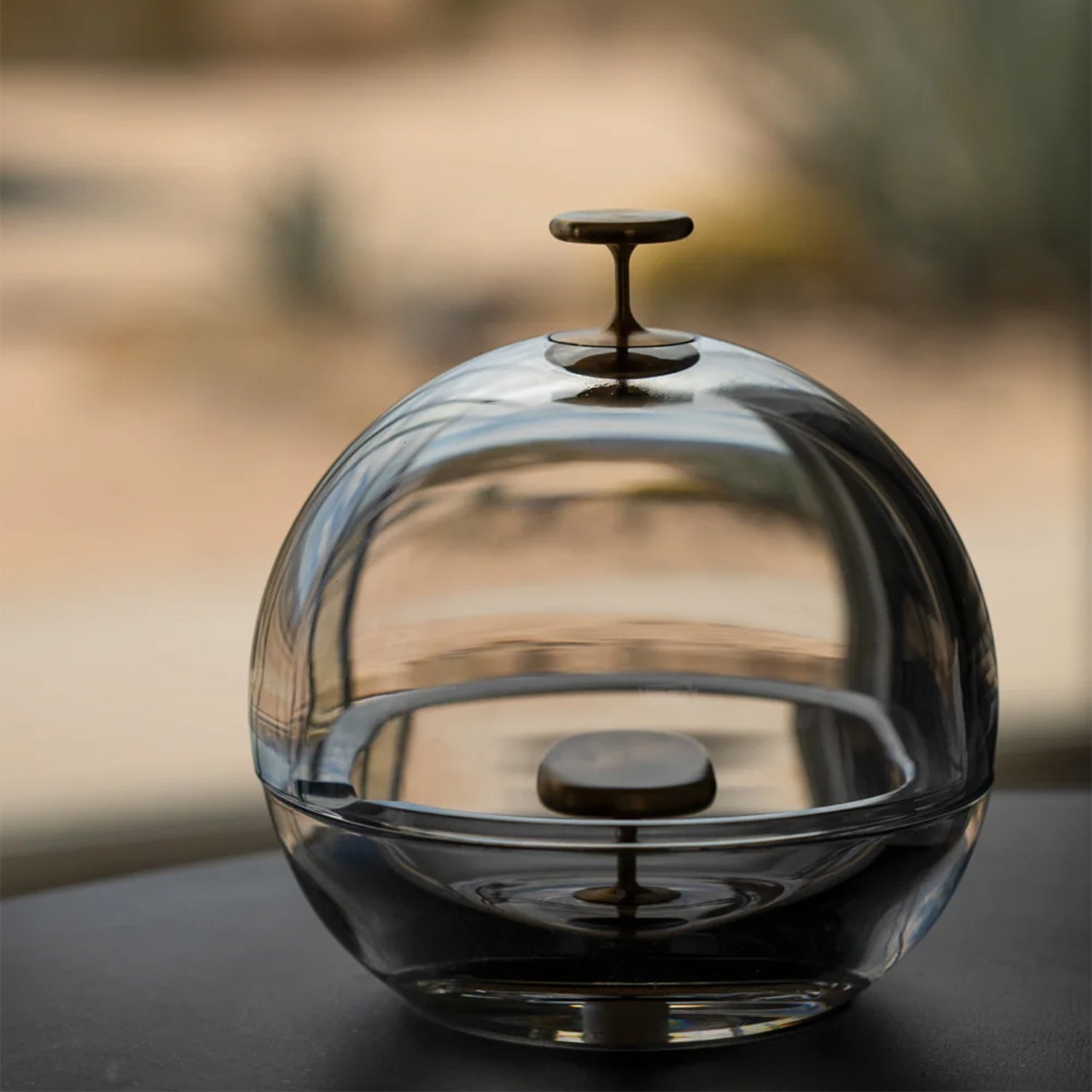 A clear glass decorative dome ashtray with a dark metal knob on top, shown on a dark surface with a softly blurred warm-toned background.