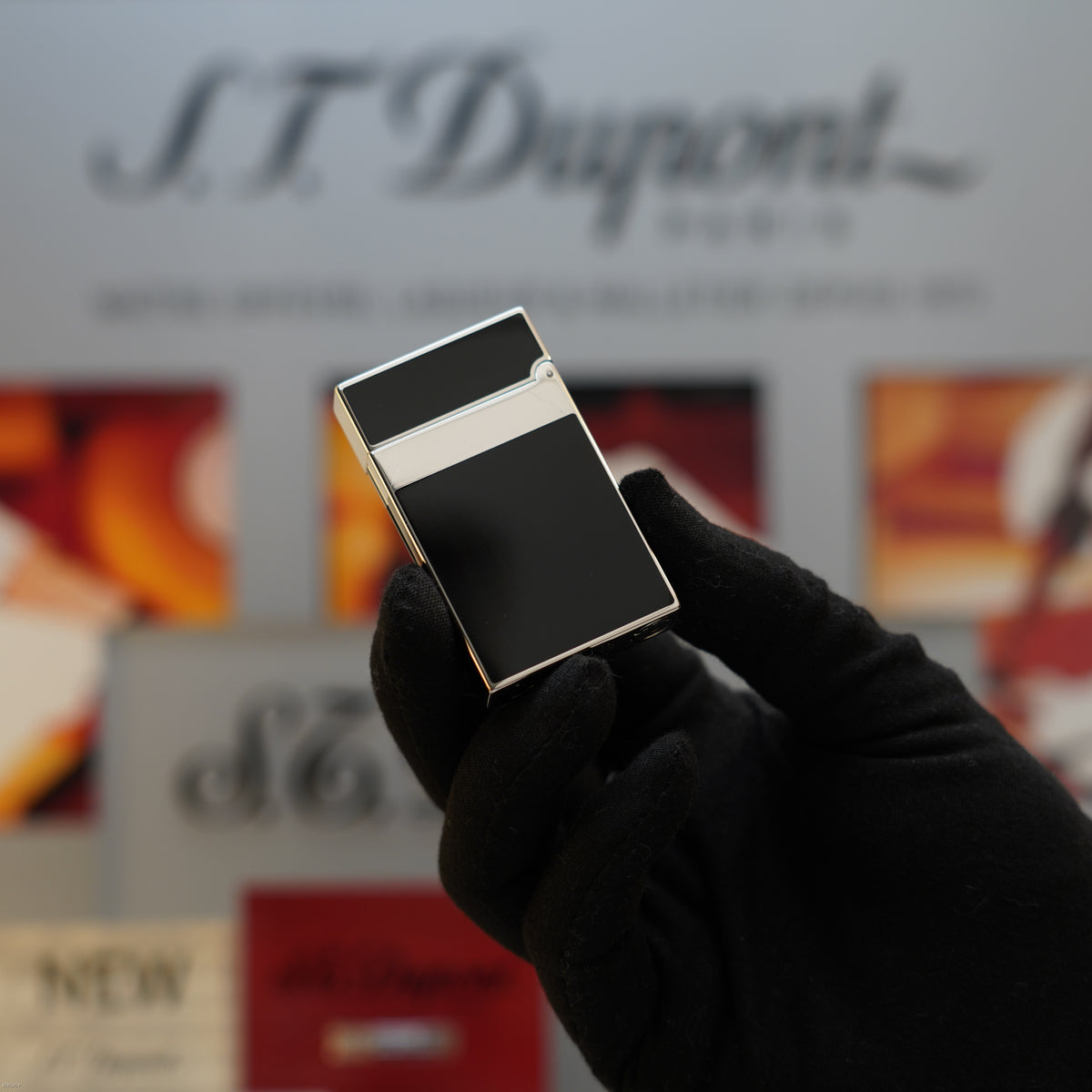 Black rectangular lighter with platinum trim and white stripe, held in gloved hand against S.T. Dupont branded background.