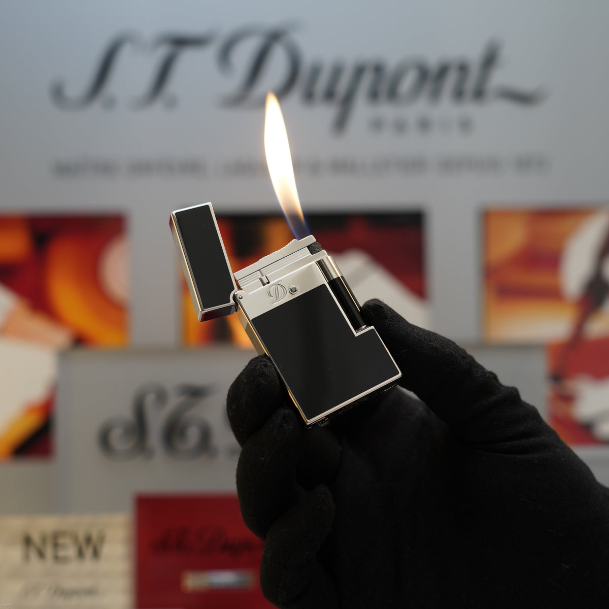 Black lacquer rectangular lighter with platinum band and lit flame, held in gloved hand against S.T. Dupont branding.