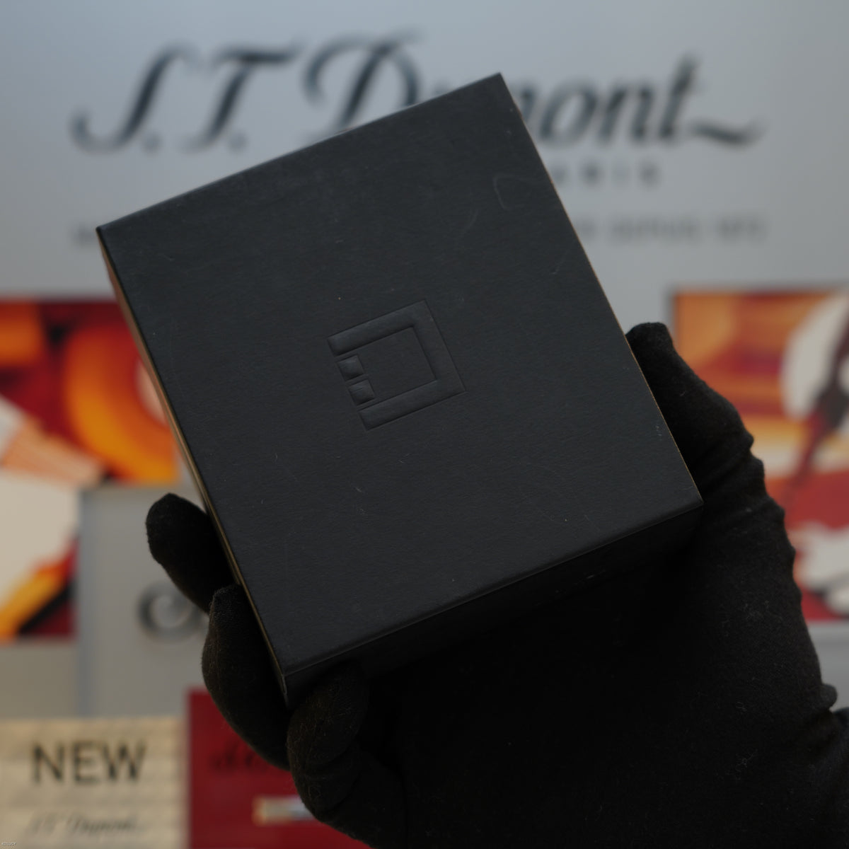 Black square lighter with embossed diamond pattern held in gloved hand, S.T. Dupont branding visible in background.