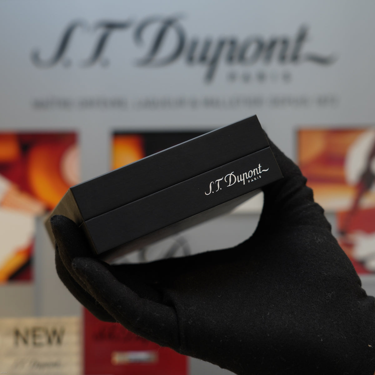 Black S.T. Dupont lighter with engraved branding held in a black gloved hand, with branded packaging visible in background.