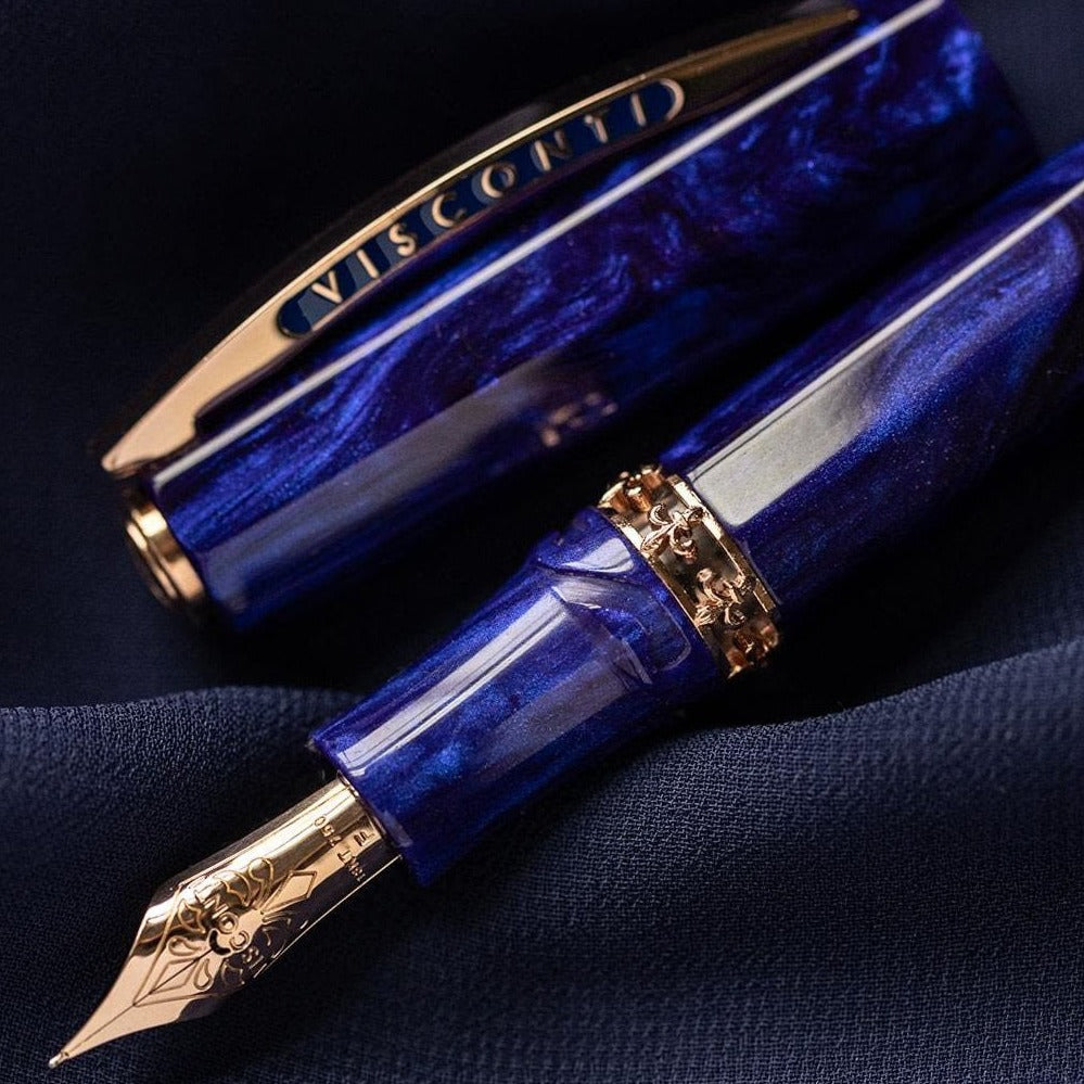 A close-up of a blue and gold Visconti Medici Viola Violet - Fountain Pen (18kt Gold Nib) resting on dark fabric. The luxury pen cap and nib are prominently visible.